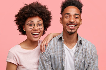 Joyful dark skinned friends being in good mood after noisy party with friends, stand closely, smile broadly, laugh at good joke. African American sister and brother pose together against pink wall