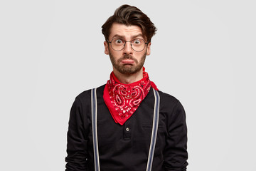 Insulted displeased European male with thick beard, curves lower lip, has trendy haircut, wears fashionable shirt with red bandana, isolated over white background. Offended guy stands indoor