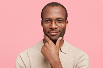 Headshot of serious dark skinned male boss holds chin, contemplates about succeessful agreement, looks mysteriously at camera, dressed in casual t shirt, stands indoor against pink background - obrazy, fototapety, plakaty