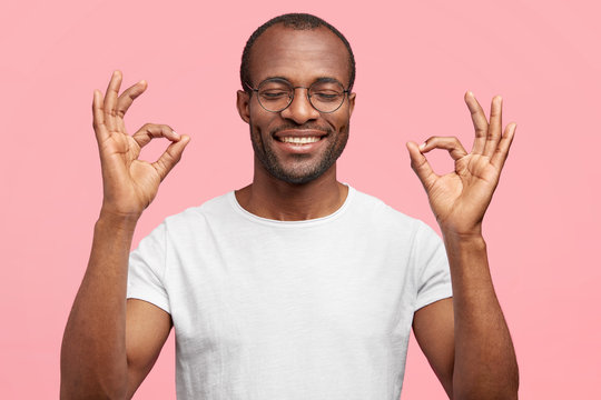 Okay, I will deal with it! Happy African American middle aged male with broad toothy smile, shows ok gesture, closes eyes in pleasure, dressed casually, isolated on pink background, approves something