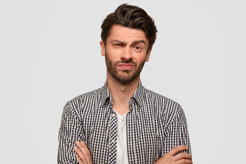 Stylish hipster male keeps arms folded, looks with hesitant expression, raises eyebrow with...
