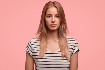 Horizontal shot of pretty Caucasian female with serious expression, healthy skin, light hair, dressed in striped t shirt, poses over pink background has appealing appearance. Student thinks about exam