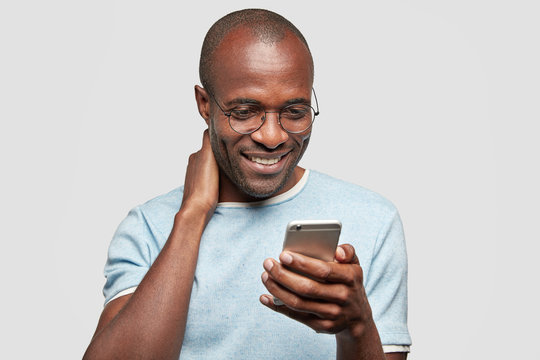 Cheerful African American guy chats on smart phone with friend or girlfriend, recieves good news in message, holds modern cell phone, wears spectacles and t shirt, isolated over white studio wall
