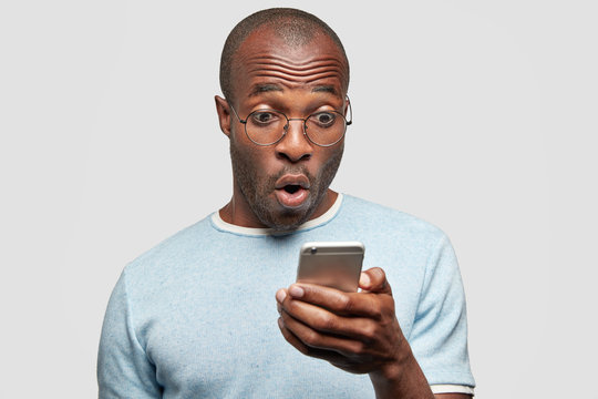 Photo of stunned African Ameriican male reads text message with surprised expression, holds cell phone, finds out something shocking, connected to wireless internet. People and technology concept
