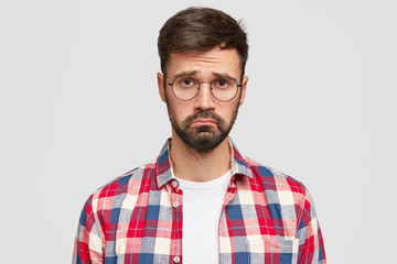 Fototapeta premium Upset young bearded male with gloomy expression, being offended by someone, raises eyebrows, needs support and help, wears glasses and casual checkered shirt, stands indoor, being in low spirit