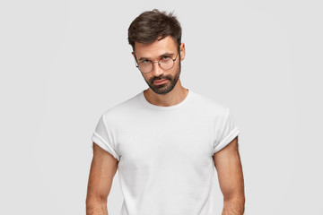 Serious bearded man with self assured expression, looks directly at camera, wears round spectacles, has calm facial expression, dressed in casual t shirt, stands alone against white background