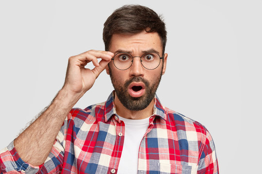 Scared puzzled Caucasian male with frightened expression, stares through glasses, opens mouth widely, holds hand on rim of spectacles, dressed in checkered shirt, isolated over white background