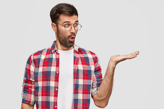 Emotional bearded male student with surprised expression keeps palm raised against white background, stares with bugged eyes, dressed casually, shows space for your advertising content or promotion
