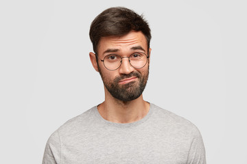 Puzzled doubtful unshaven male curves lips, frowns face with hesitation, has clueless expression, wears casual grey clothes, isolated over white background. People, reaction, facial expressions