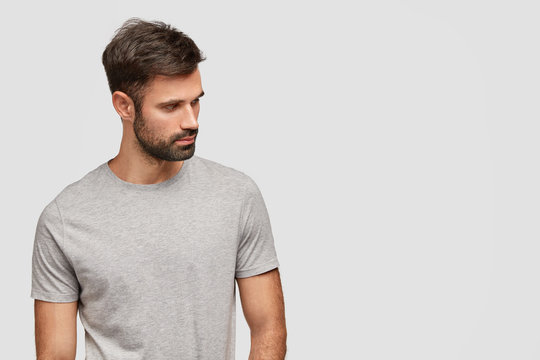 Indoor shot of concentrated handsome unshaven young male foused aside, has muscular body, dressed in casual grey t shirt, stands against white background with blank space for your advertisement