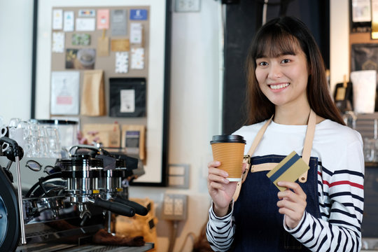 Young asian woman barista holding a disposable coffee cup and credit card with smiling face at cafe counter background, small business owner, food and drink industry concept