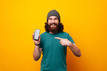 Cheerful bearded hipster man smiling and poiting at smartphone over yellow background