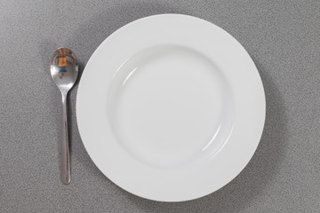 Suppenlöffel aus Metall neben einem weißen Suppenteller auf einem grauen Tisch 