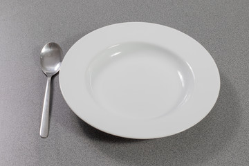 Suppenlöffel aus Metall neben einem weißen Suppenteller auf einem grauen Tisch 