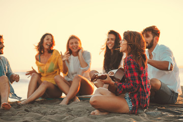 Happy friends sitting on the beach singing and playing guitar during the sunset