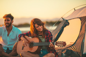 Happy friends sitting on the beach singing and playing guitar during the sunset