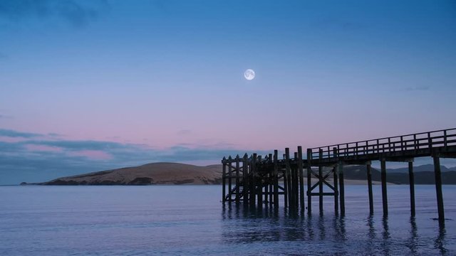 View across calm lapping water in Hokianga Harbour at dawn with full moon rising over wooden pier. New Zealand.