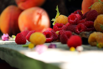 Still life with red and yellow raspberries and peaches. Fruit on a wooden table. Against the background of a bouquet of wildflowers. Close-up. Selective focus, side view, copy space.