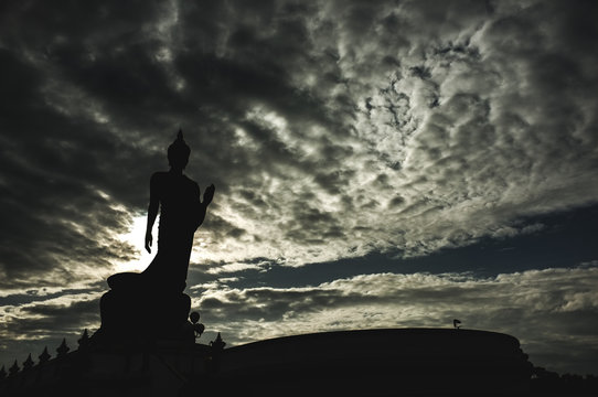 Stand big tall Buddha Statue With sun set Light background in public park of thailand temple. Buddha image represent the Lord Buddha. Sky with black clouds - gold as background.