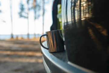 Closeup iron camping mug with a warm drink on the shelf with the open door of the rear trunk of the SUV on the camping background. Travel by SUV or car in the wild. Concept of camping in the forest.