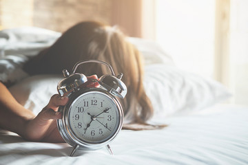 Irritated young woman putting her alarm clock off in the morning with soft morning light. Relaxing concept. - obrazy, fototapety, plakaty