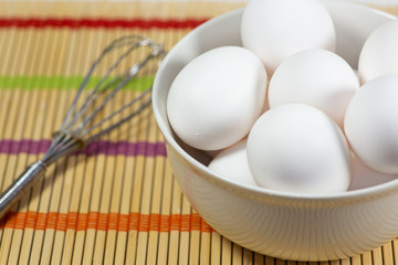  A group of eggs inside a deep white bowl next to a whisk waiting for the chef to use them in a meal on a bamboo mat