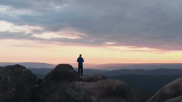 Drone shot of lonely man standing on top of a mountain and enjoying the sunset