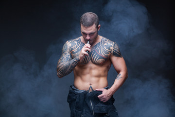 Fototapeta na wymiar Vaper. The man with a muscular naked torso with tattoos smoke an electronic cigarette on the dark background