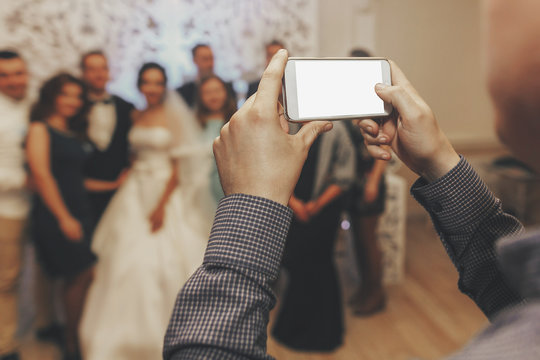 hand holding phone with empty screen and taking photo of happy wedding couple and guests at wedding reception.photo booth for bride and groom in restaurant.