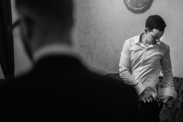 Groom getting ready in the morning with groomsmen in the room. man hands putting on jacket and bow tie, standing at window light in hotel. preparation for wedding day