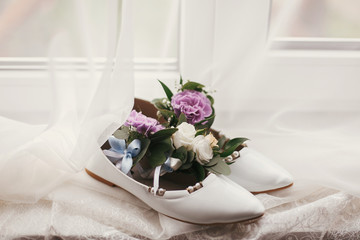 stylish white shoes with flowers in them on window in soft morning light. creative image. beautiful bridal getting ready for wedding day. bridal shoes with bouquet