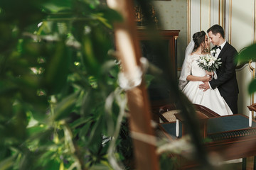 gorgeous bride with bouquet and stylish groom gently hugging among green leaves in luxury room in hotel. rich wedding couple embracing. sensual moment of newlyweds in classic indoors