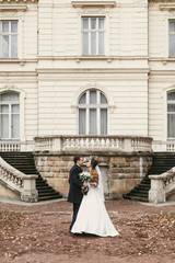 Obraz premium gorgeous bride with bouquet and stylish groom walking at old castle in european city street in autumn time. happy wedding couple embracing. romantic sensual moment of newlyweds