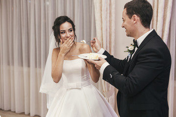 gorgeous bride and stylish groom tasting their stylish wedding cake at wedding reception in...