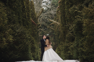 gorgeous wedding couple kissing in winter snowy park. stylish bride in coat and  groom embracing...