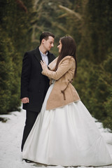Fototapeta premium gorgeous wedding couple posing in winter snowy park. stylish bride in coat and groom embracing under green trees in winter forest. romantic sensual moment of newlyweds