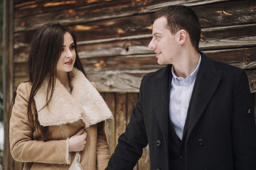 gorgeous bride in coat and stylish groom posing at wooden house in winter forest. happy wedding couple gently hugging in snowy park.  barn wedding. romantic sensual moment