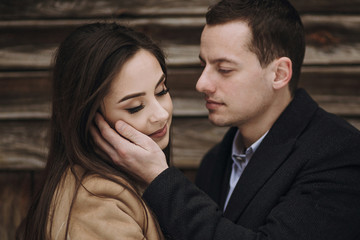 gorgeous bride in coat and stylish groom hugging at wooden house. wedding couple gently hugging and embracing in snowy park. barn wedding. romantic sensual moment of newlyweds