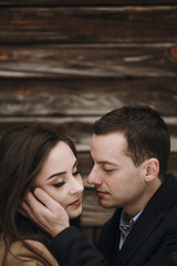 gorgeous bride in coat and stylish groom hugging at wooden house. wedding couple gently hugging and embracing in snowy park. barn wedding. romantic sensual moment of newlyweds