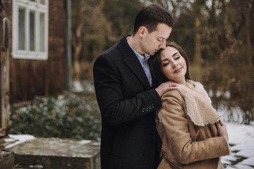 gorgeous bride in coat and stylish groom posing at wooden house in winter forest. happy wedding couple gently hugging in snowy park.  barn wedding. romantic sensual moment