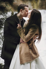 gorgeous wedding couple kissing in winter snowy park. stylish bride in coat and  groom embracing under green trees in winter forest. romantic sensual moment of newlyweds