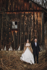 gorgeous bride in coat and stylish groom posing at wooden house. wedding couple gently hugging and...