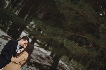 gorgeous wedding couple posing in winter snowy park. stylish bride in coat and  groom embracing under green trees in winter forest. romantic sensual moment of newlyweds