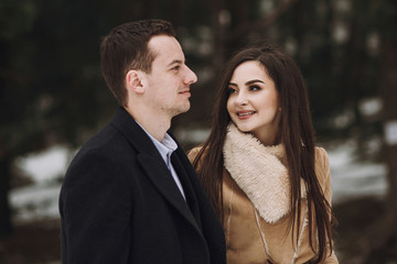 Fototapeta premium gorgeous wedding couple walking in winter snowy park. stylish bride in coat and groom embracing under green trees in winter forest. romantic sensual moment of newlyweds