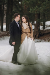 Fototapeta premium gorgeous wedding couple walking in winter snowy park. stylish bride in coat and groom embracing under green trees in winter forest. romantic sensual moment of newlyweds