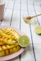Grilled pineapple slices on a pink plate with honey and lime over striped napkin, side view. Summer food. Closeup. Selective focus.