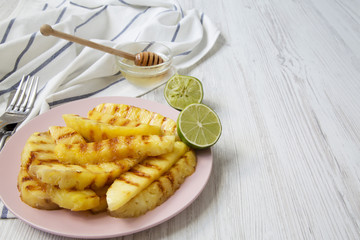 Grilled pineapple wedges on a pink plate with honey and lime over white wooden background, side view. Summer food. Closeup. Copy space.