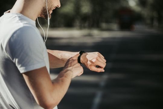 Focus on smiling man standing and looking at fitness tracker on wrist. He is jogging with music on his own with delight