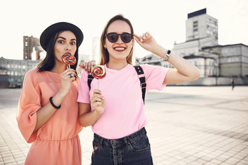 Portrait of beaming lady touching sunglasses while keeping candy in hand. She standing near glad...
