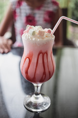 Strawberry milkshake in a glass, with marshmallows and whipped cream and a red straw. restaurant table